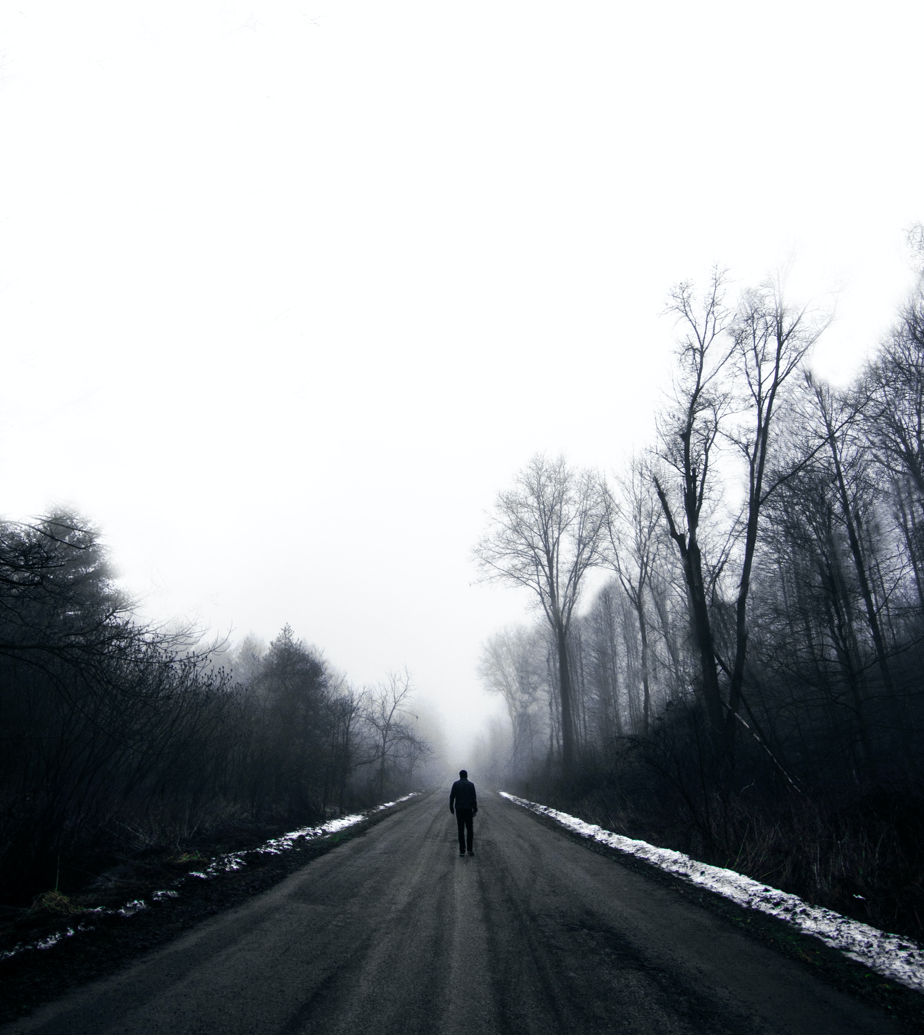 A black and white dreary winter scene of a person walking alone down an empty road away from the viewer into the distance