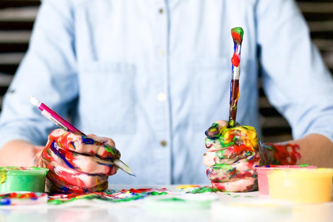 a painter's paint covered hands at work on a canvas, holding brushes in both hands.