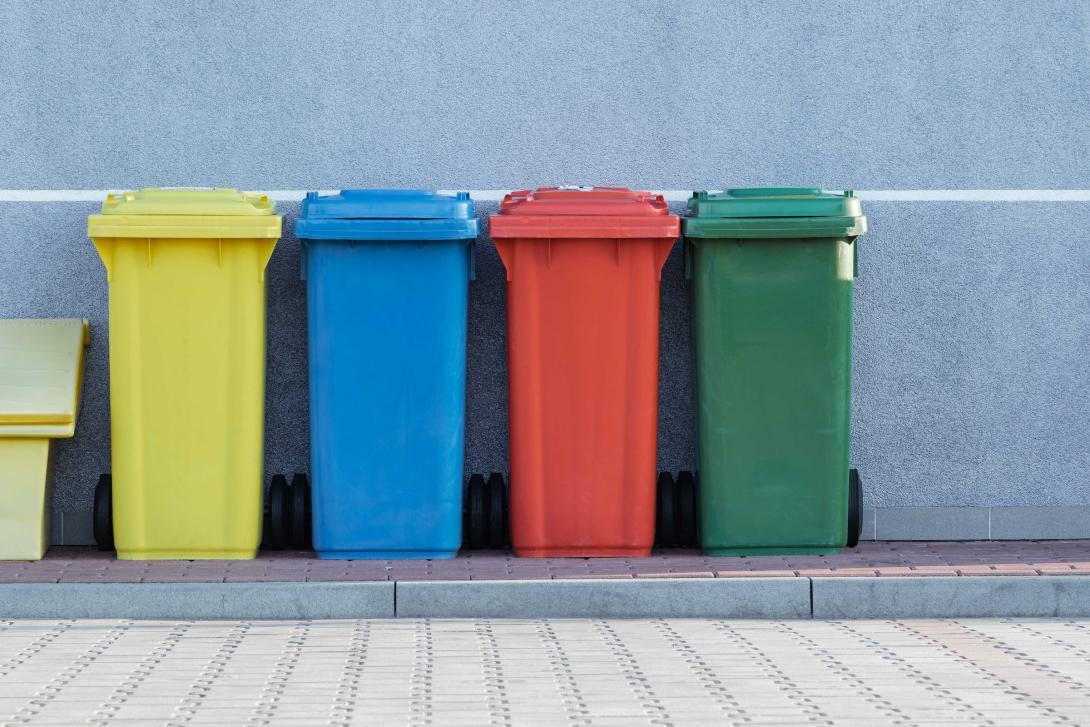 A picture of a rainbow set of what appears to be recycling bins in an alley. They are brightly colored and the scene is clean and daytime.
