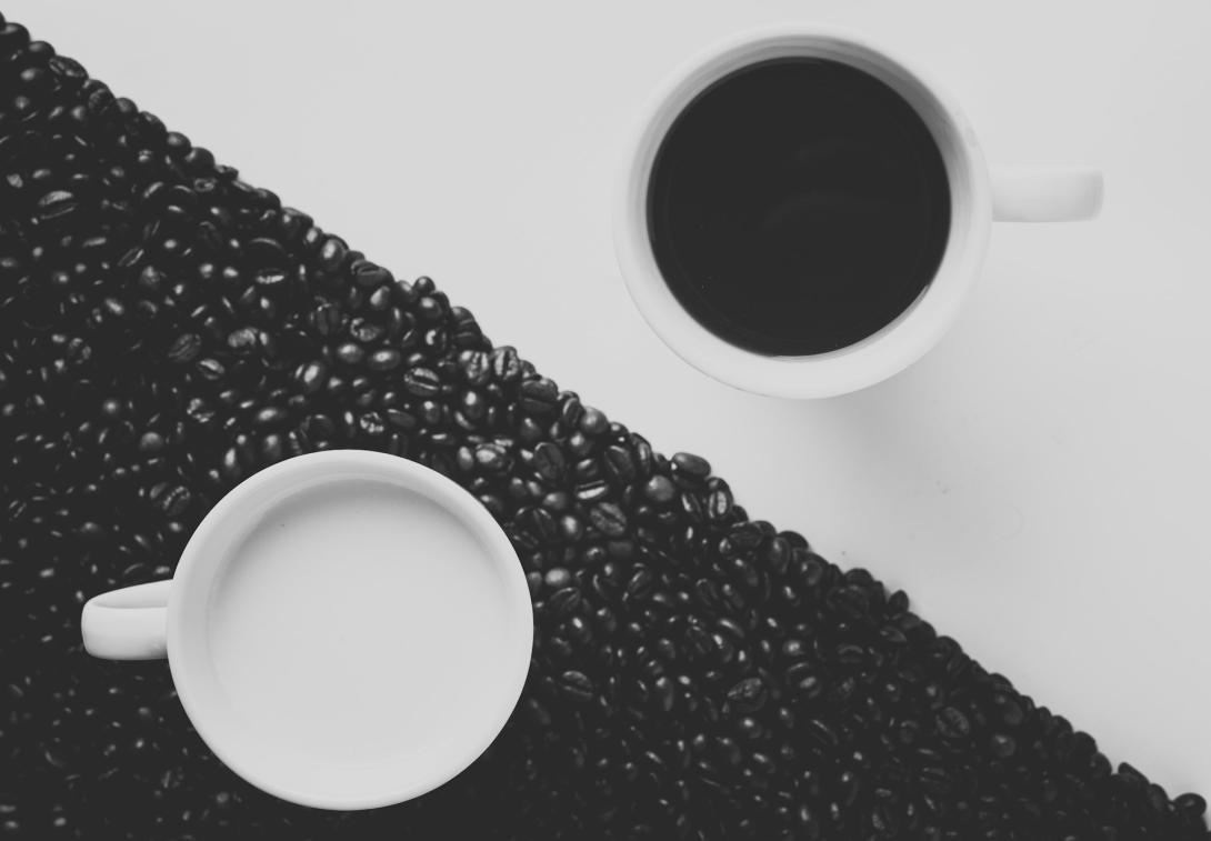 A top down picture of two coffee cups on a diagonally divided background. On the top left is a white area with a cup of black coffee, and on the bottom left is an area covered in roasted but unground coffee beans with a cup of white liquid (probably milk). The image is similar to a traditional ying-yang symbol